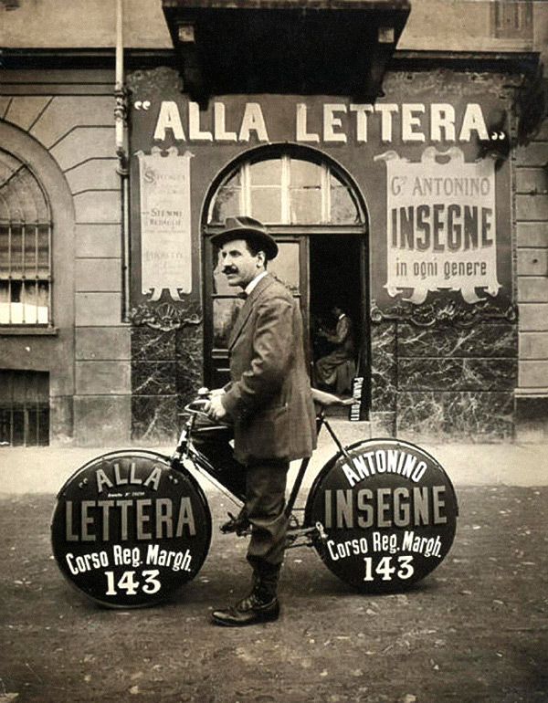 Man posing on a bike advertising his sign painting business, in front of his sign painting shop.