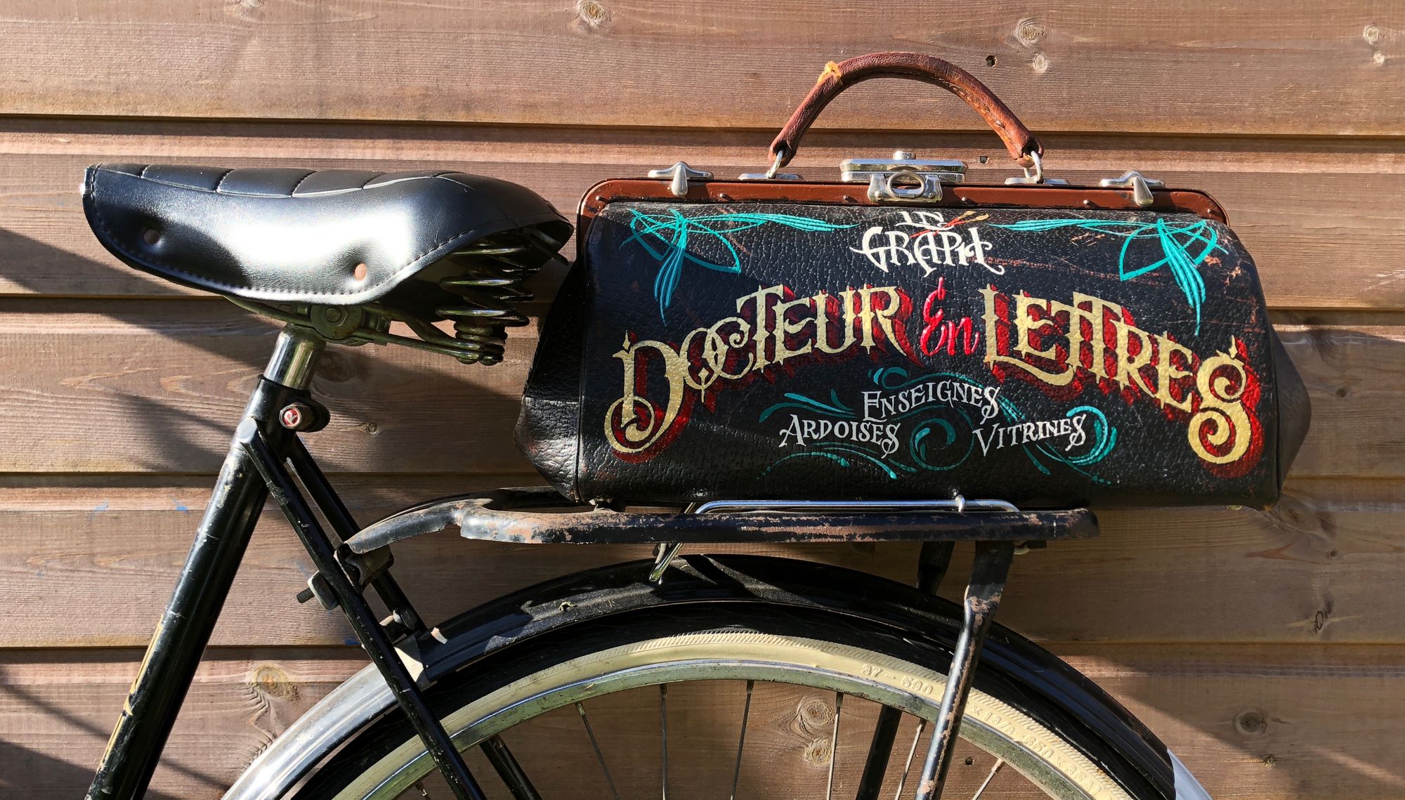 A doctor's kit bag painted to advertise a sign painter and mounted to the rack of a bike
