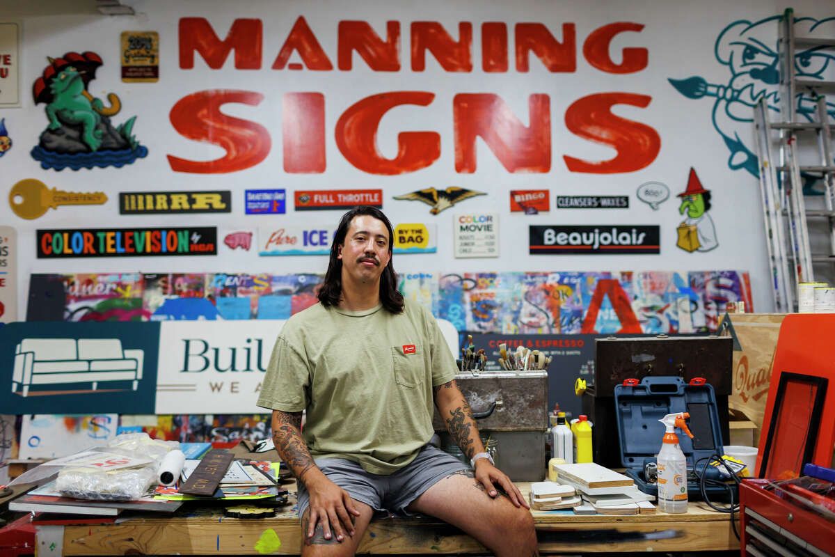 Man sitting on a workbench surrounded by a variety of painted signs.