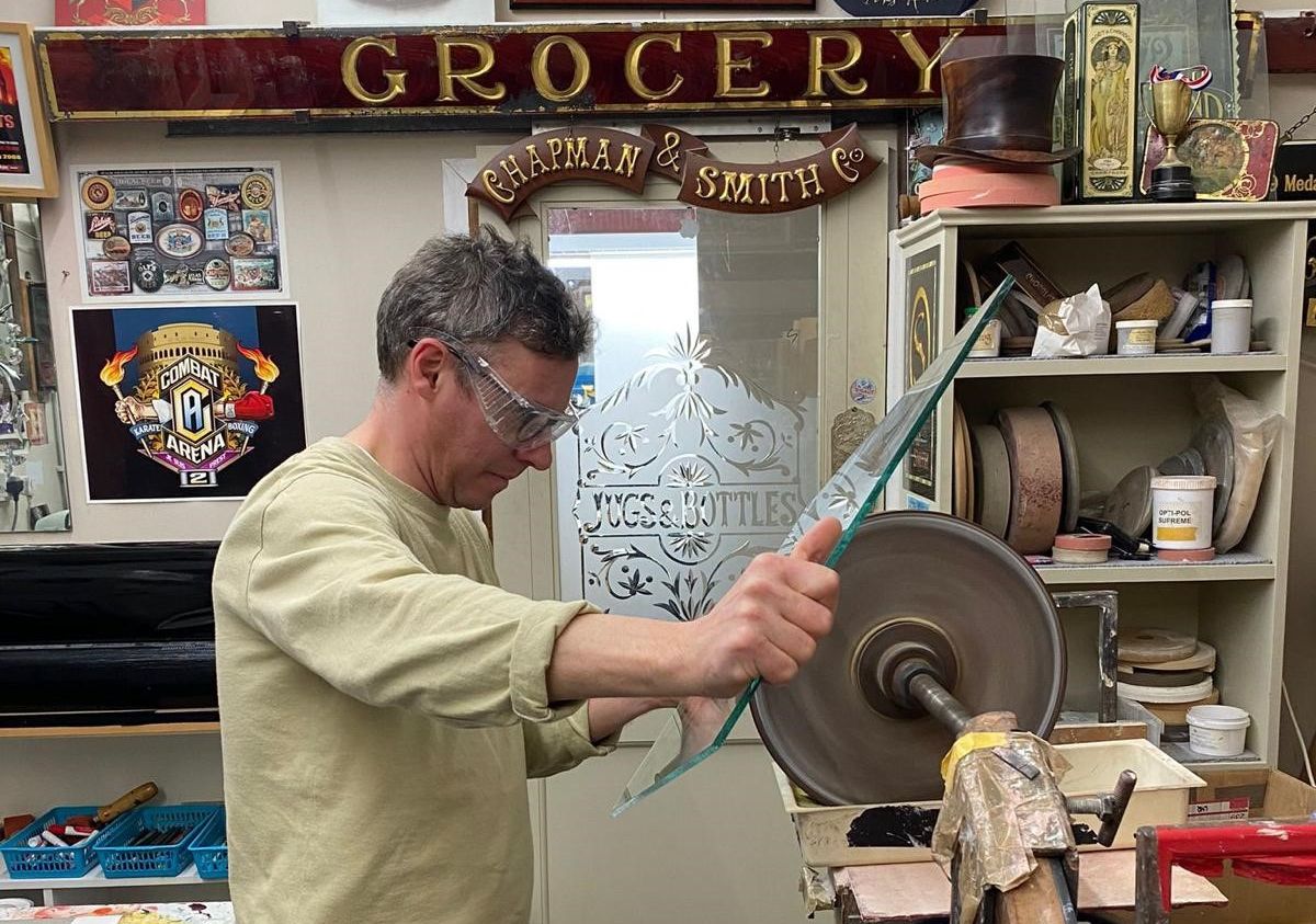 Man in protective goggles making cuts in glass with a spinning stone wheel.