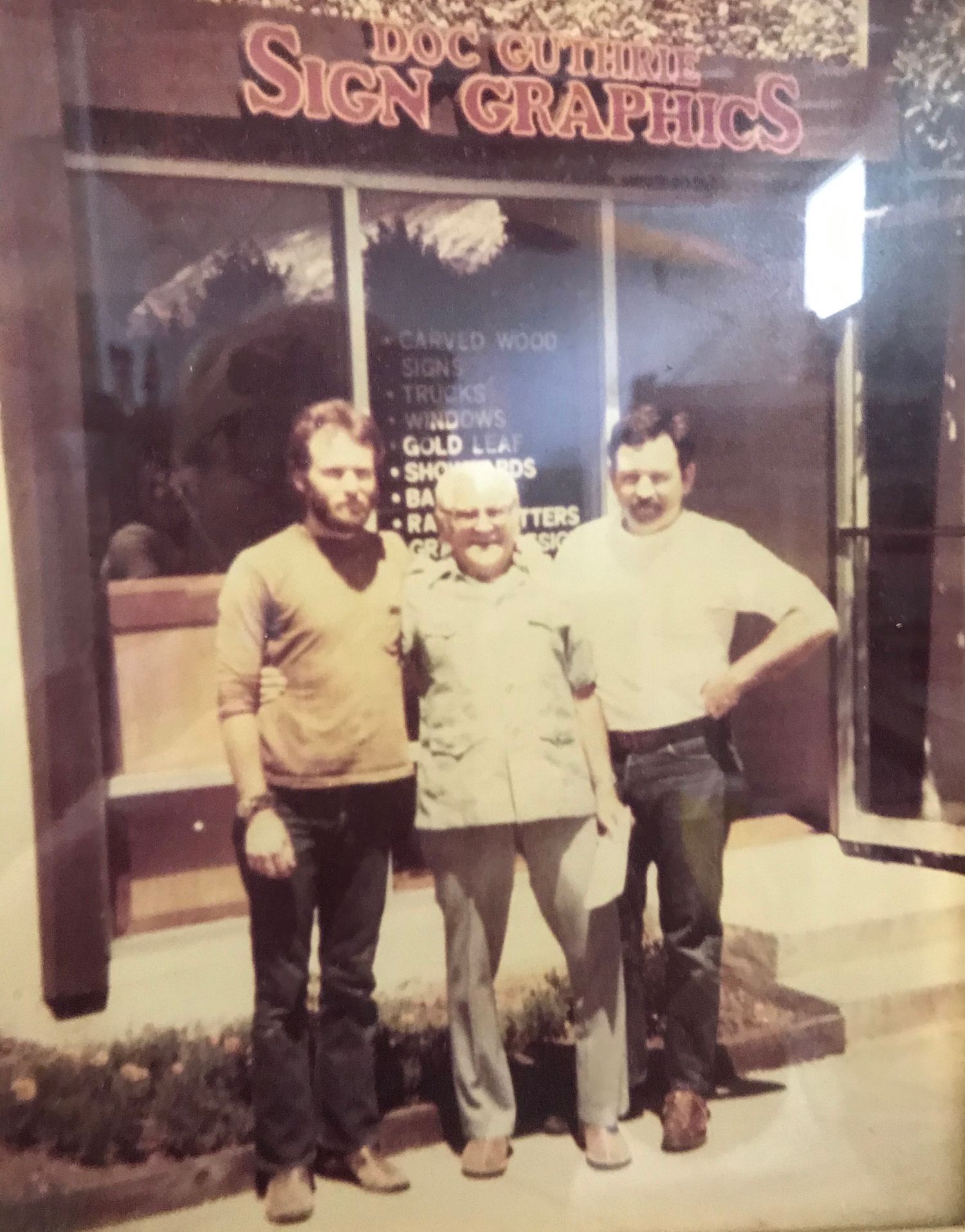 Three men standing in front of a sign shop.