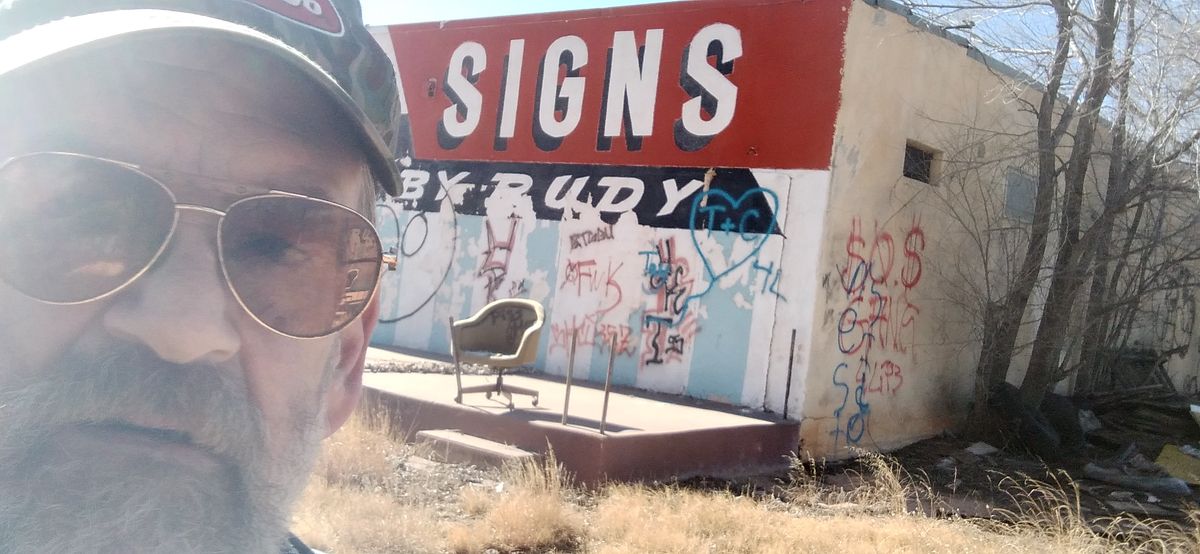 Man in sunglasses in front of an abandoned building lettered with 'Signs by Rudy'.