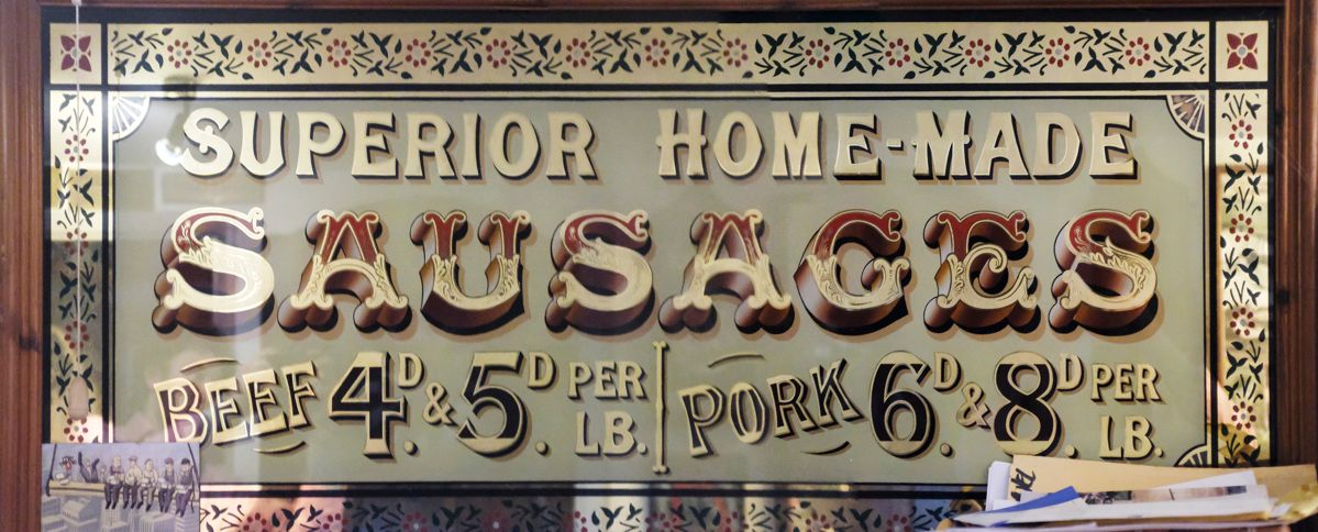Glass sign panel with painted and gilded lettering that reads "Superior Home-Made Sausages" and gives prices for beef and pork.