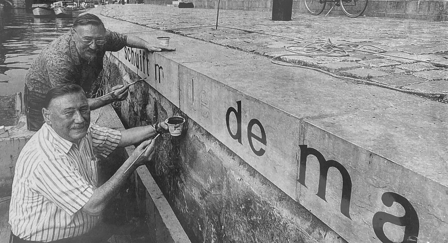 Two men posing by wall they are painting letters onto.