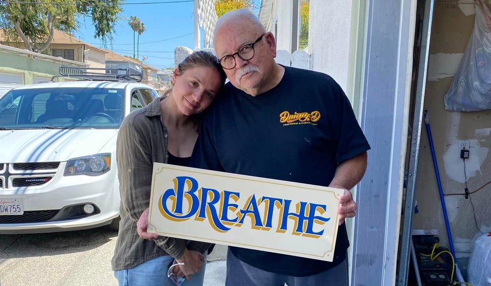 Woman and man posing with a hand-painted sign that reads 'Breathe'.