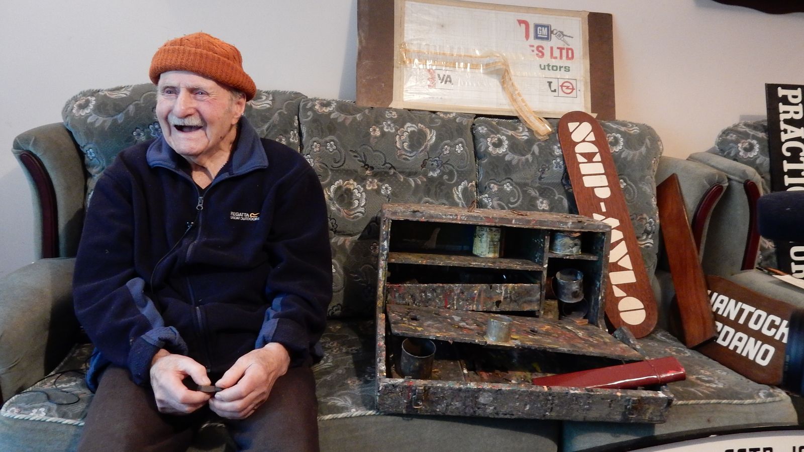 Elderly man on a sofa with his sign painting kit and various painted signs.