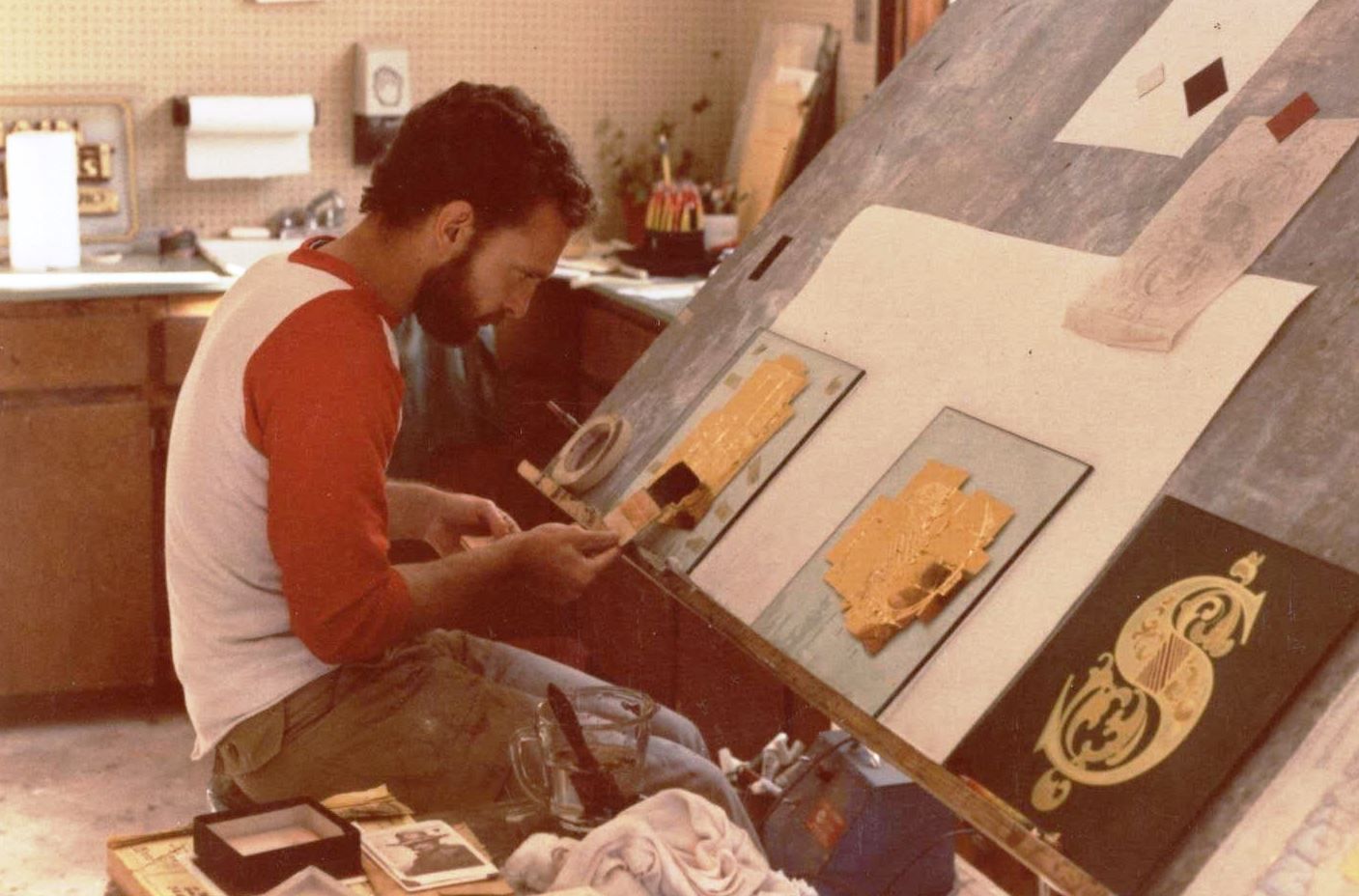 Man sat at an easel layout gold leaf onto glass panels.