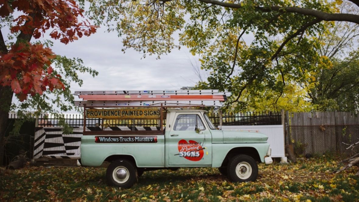 Truck with hand-painted signs on the side and ladders on the roof, parked in an autumn scene.