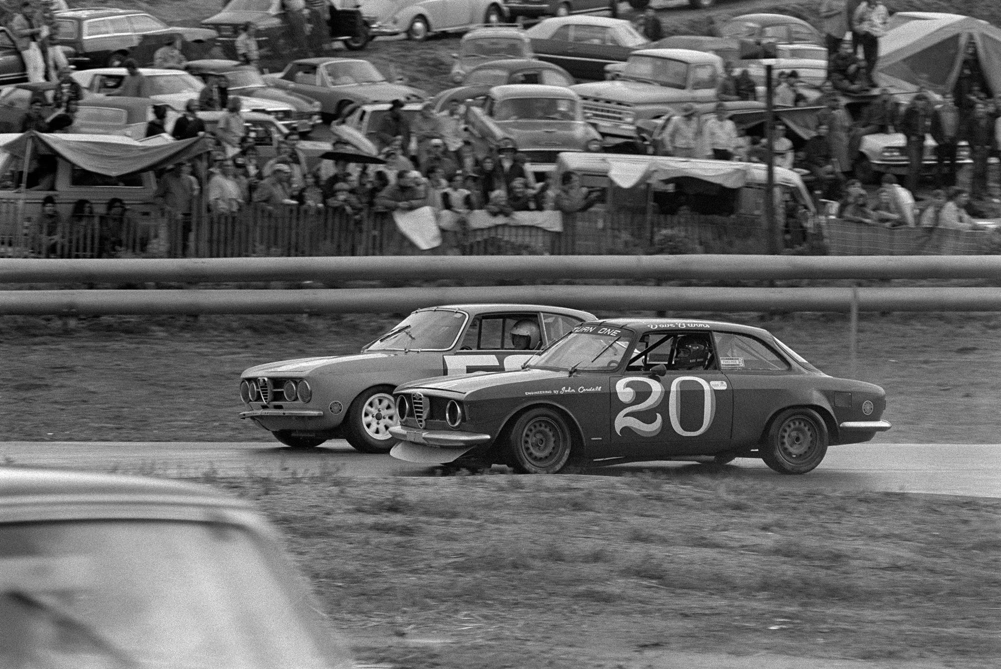 Black and white photo of two cars neck and neck in a race with a crowd looking on.