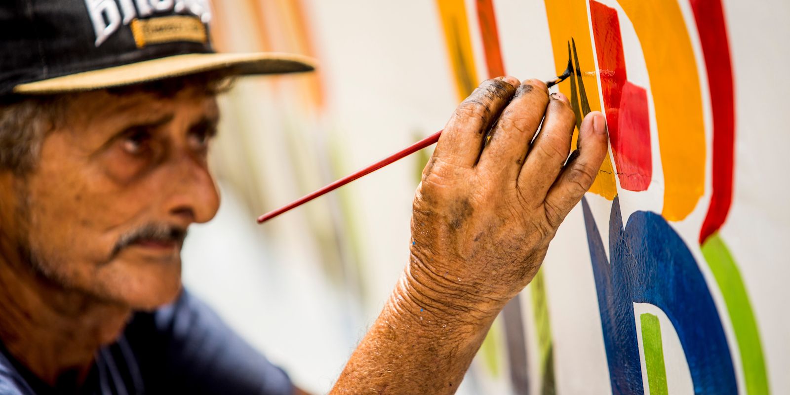 Close-up of a man in a baseball cap painting a brightly coloured letter on the side of a boat.