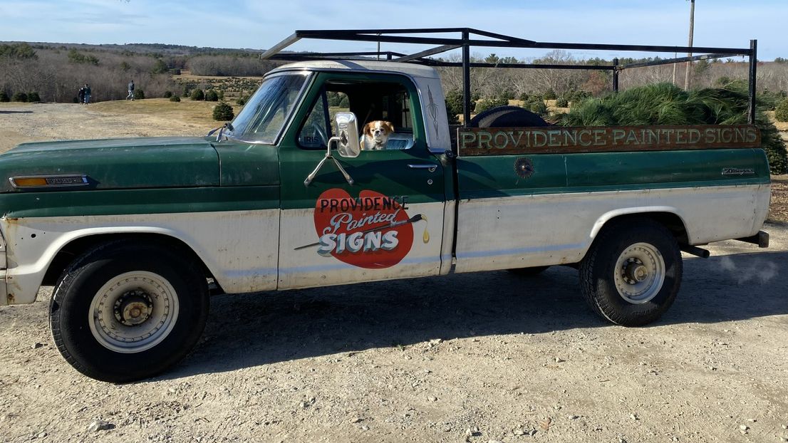 Truck with hand-painted lettering on the door and a dog in the driver's seat looking out of the window.