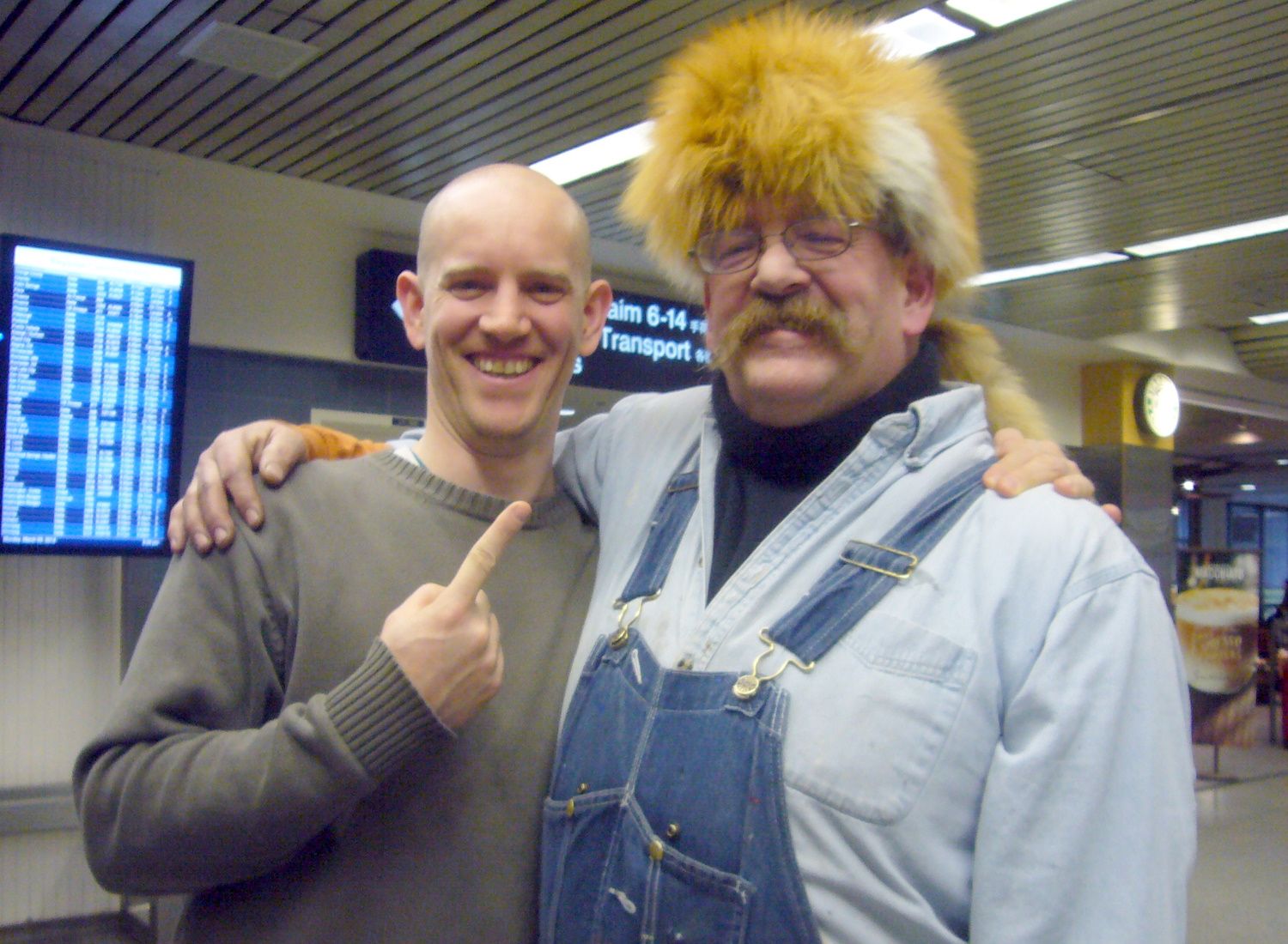 Two men standing in an airport, one wearing a fox-fur hat and the other pointing at it.