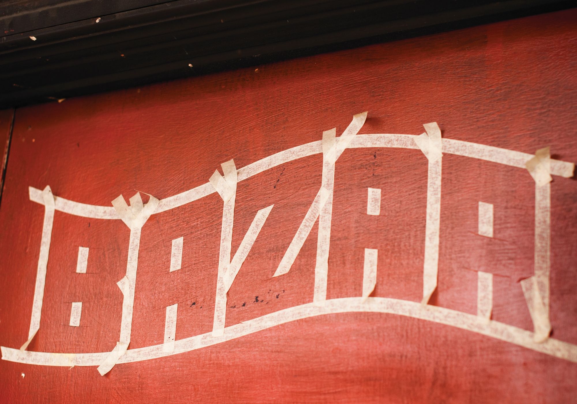 Red wall with the word 'BAZAR' outlined in masking tape ahead of painting in the letters.