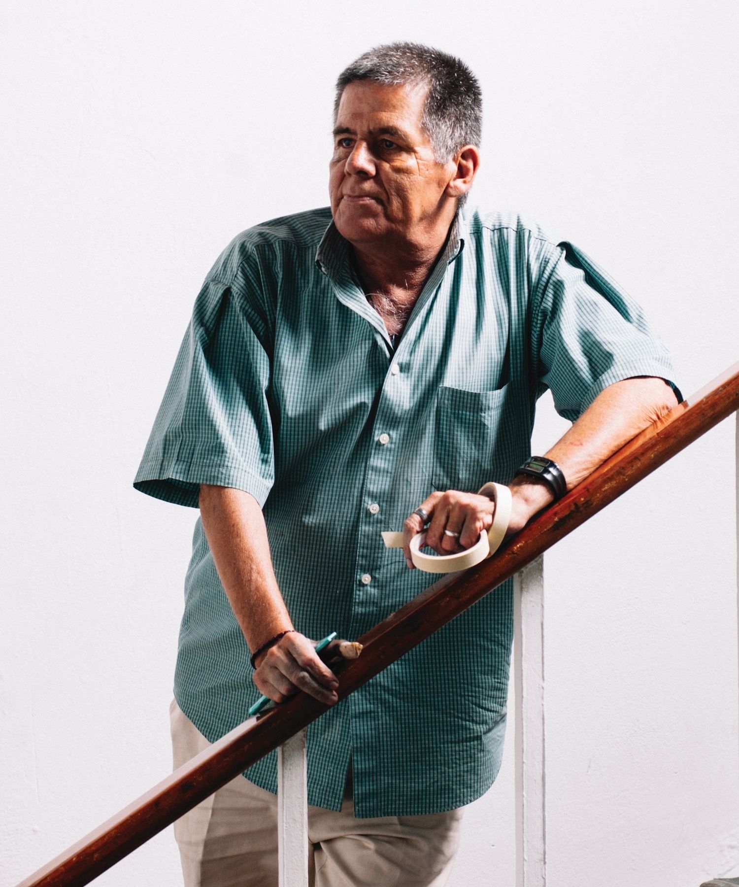Portrait of a man resting on a staircase bannister holding a pencil in one hand and rolls of masking tape in the other.