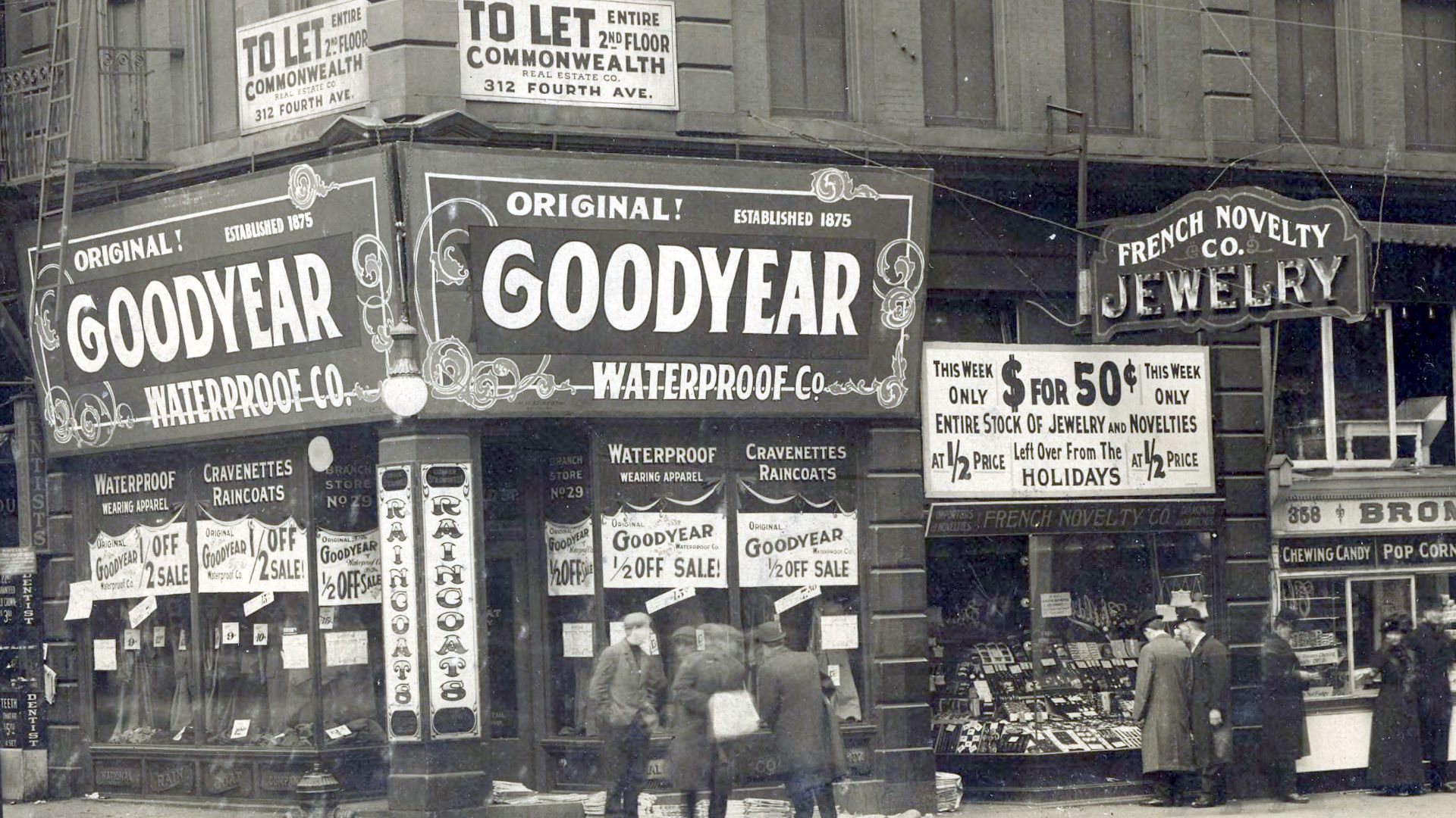 Archival image of people, shops, and signs, dominated by the corner building which houses the Goodyear Waterproof Co.