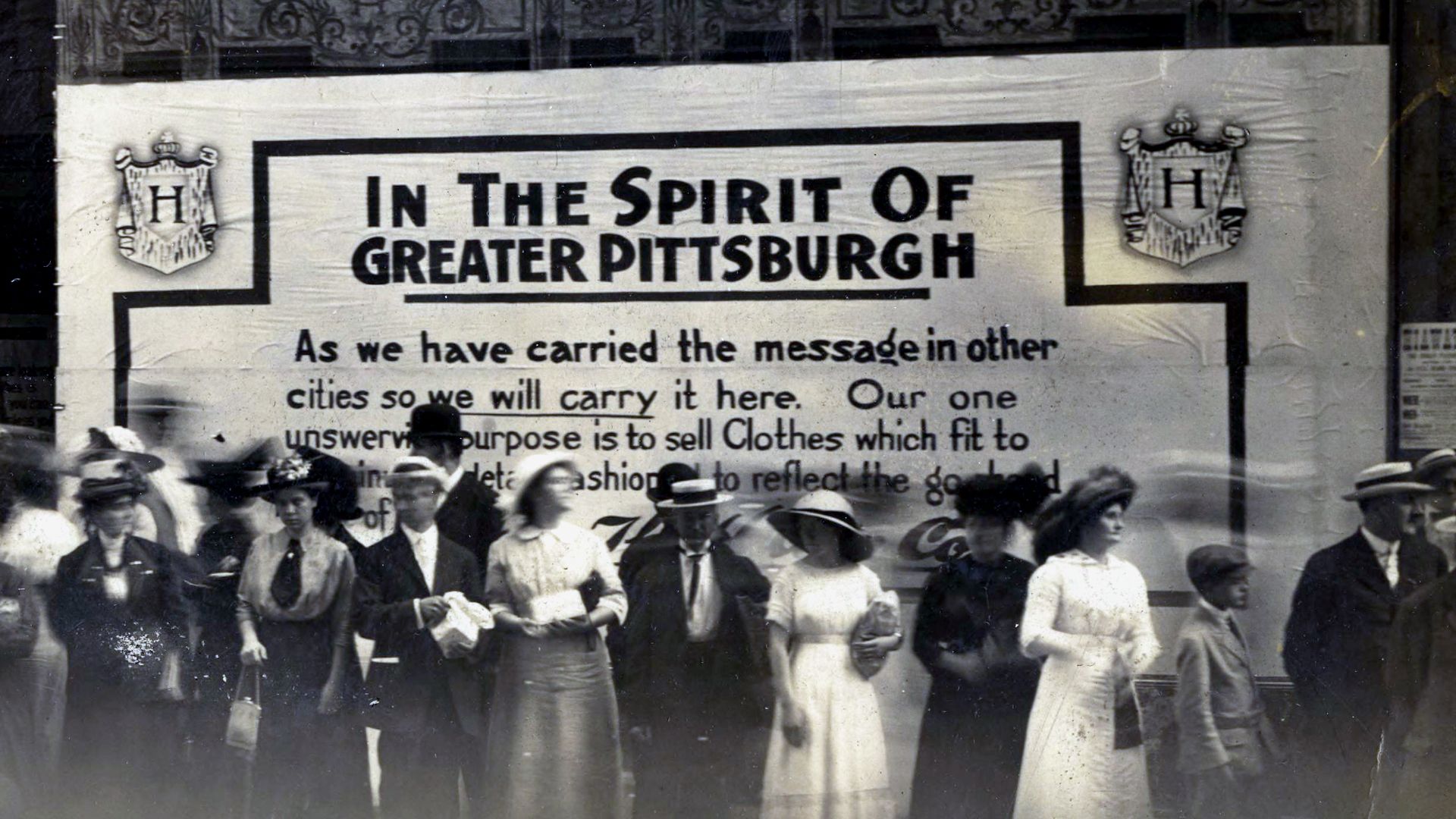 Close up of people standing in front of a painted sign announcing the opening of a new shop "in the spirit of greater Pittsburgh".
