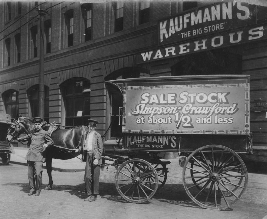 Drivers and a horse-drawn wagon adorned with temporary signage in front of a large commercial building.