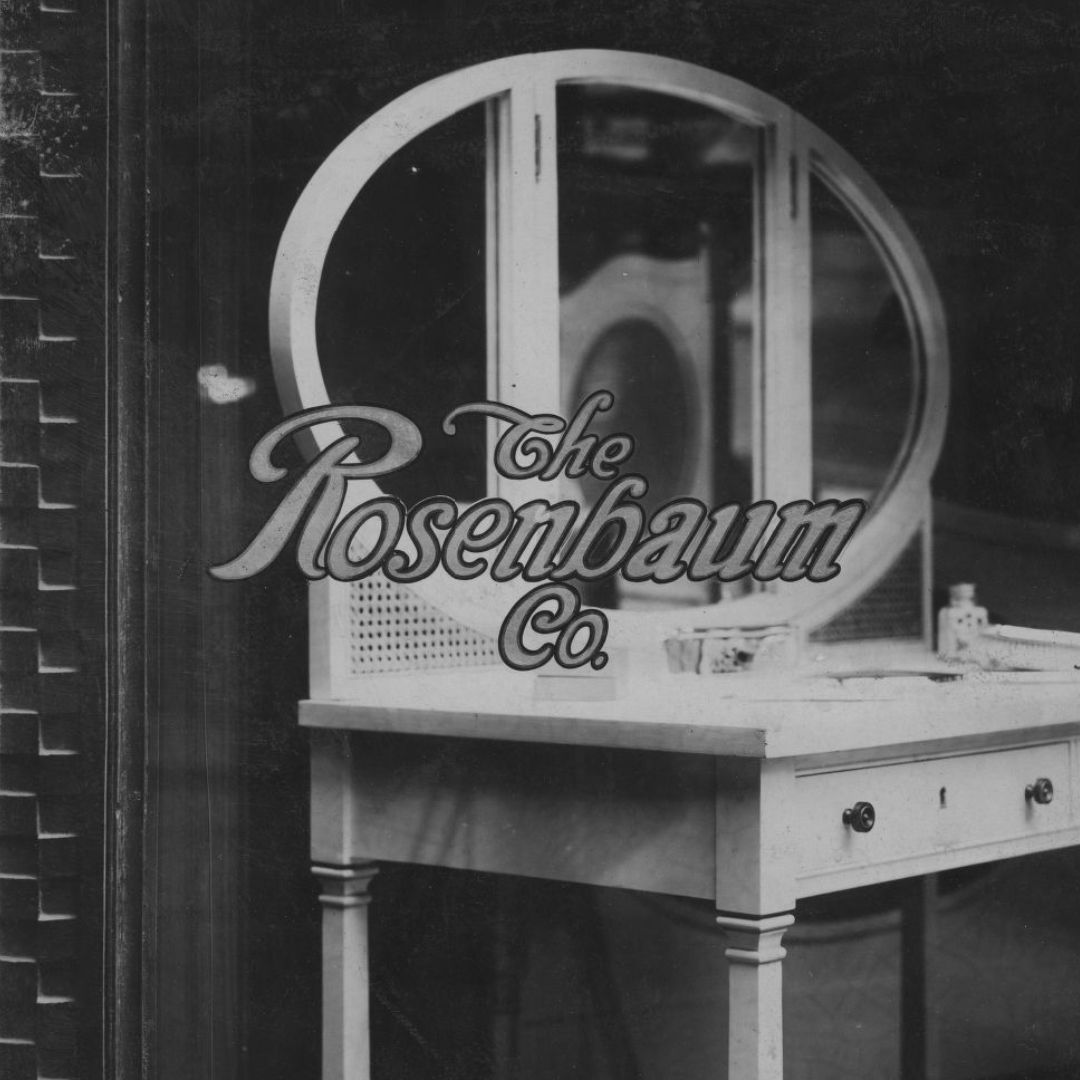 View through a shop window at a dresser with "The Rosenbaum Co." in gilded cursive lettering on the glass.