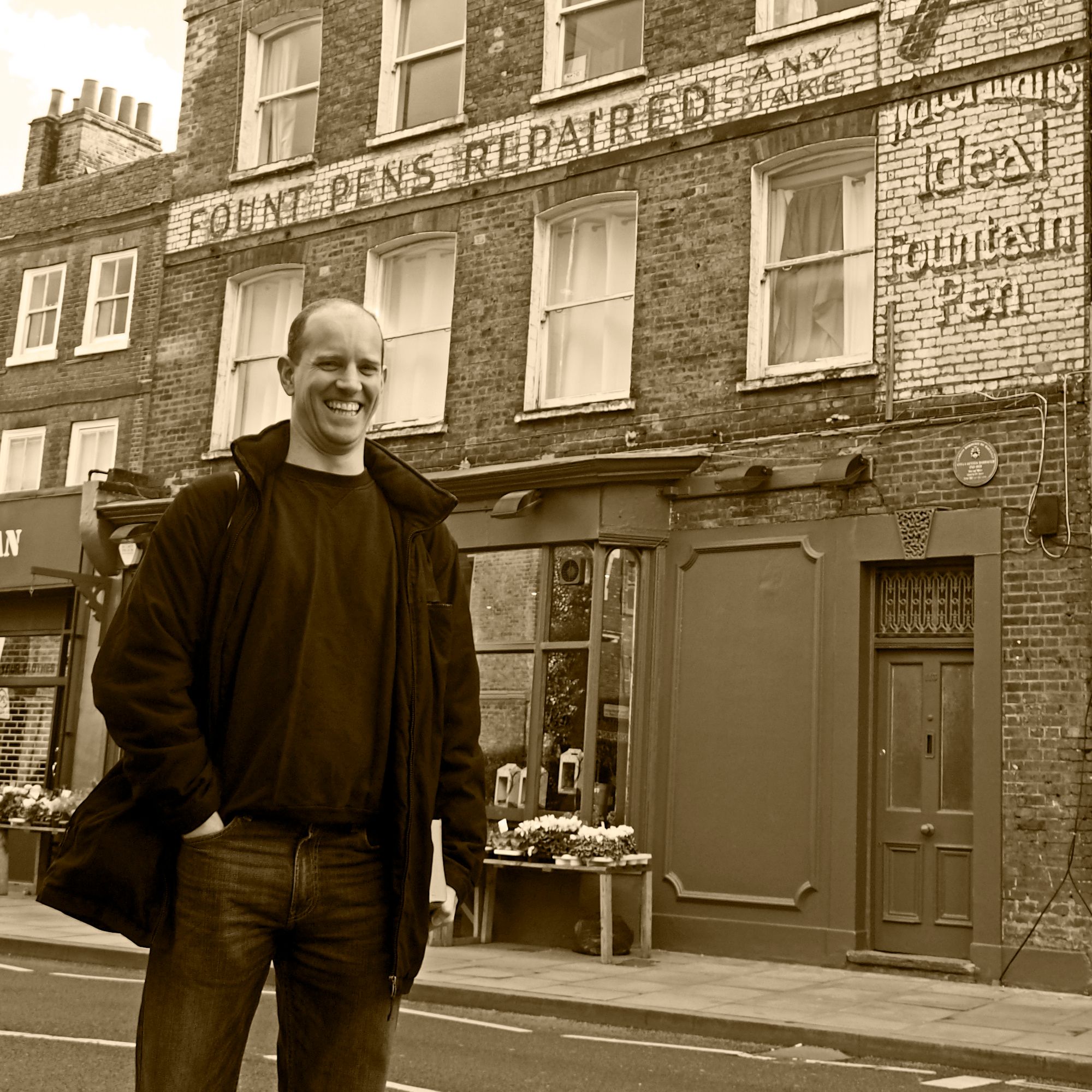 Sepia photo of a man smiling in front of a building with a hand-painted advertising sign on the brick.