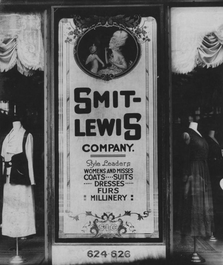 Ornate glass panel sitting between window displays of women's clothing. The piece combines lettering, pictoral and decorative elements.