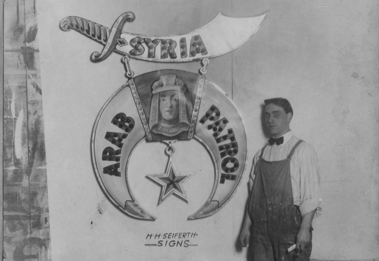 Man in overalls standing in front of a sign painted on a wall.