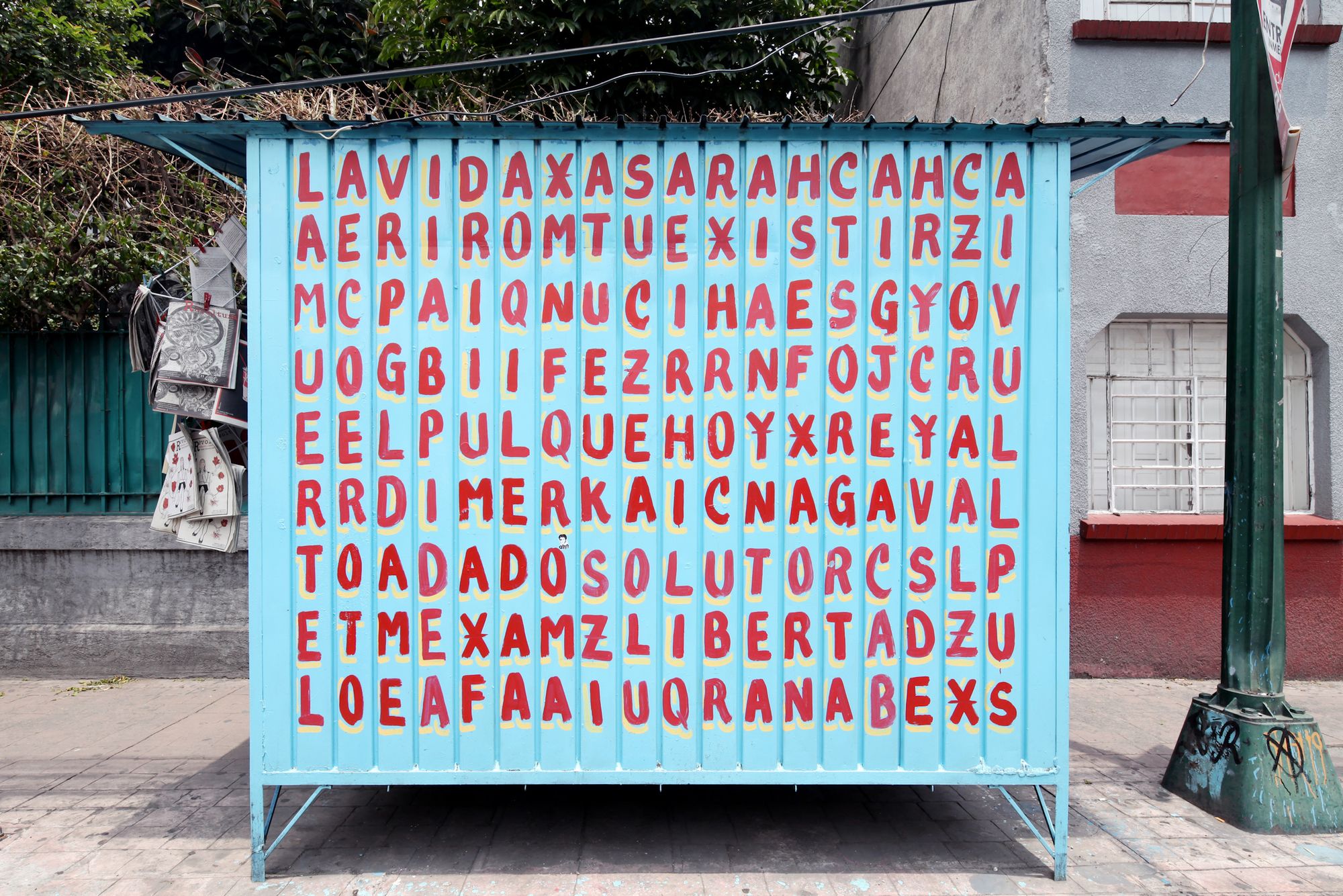 Street stand with a grid of letters painted on the light blue wooden slats in red with yellow shades.
