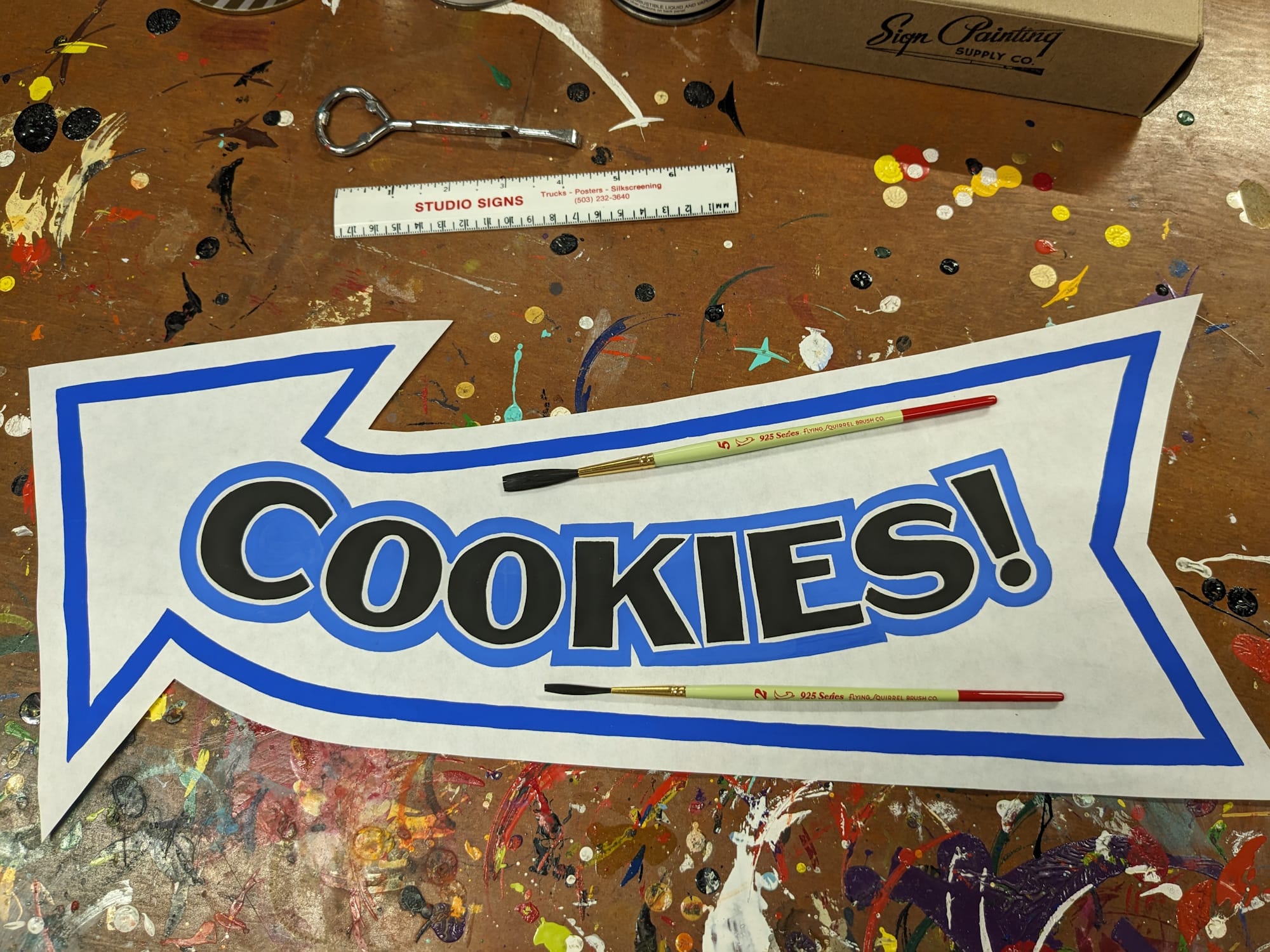 Hand-painted paper sign in the shape of an arrow with the word 'cookies' lettered in black with a blue outline.