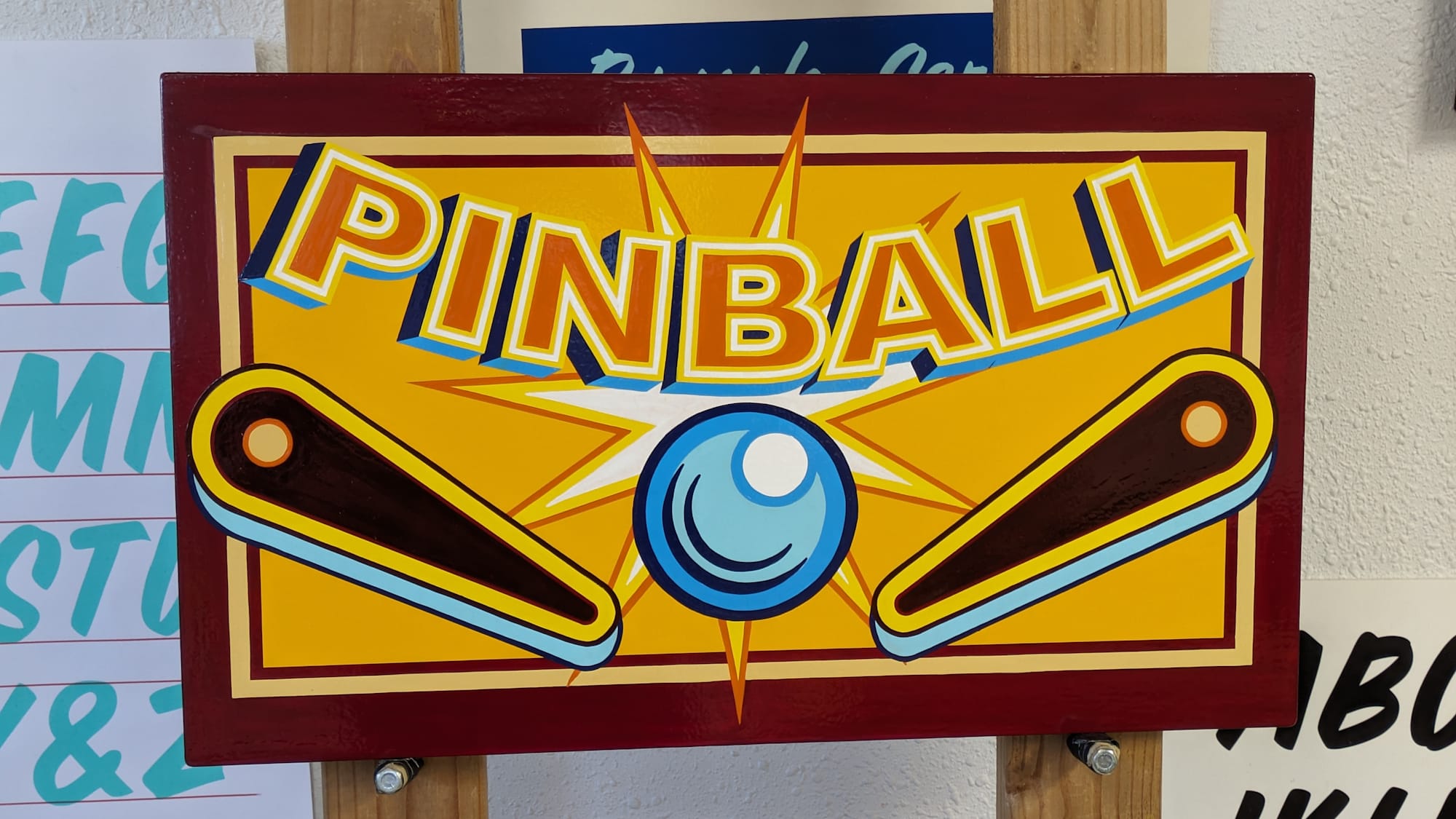 A hand-painted sign on an easel with the word 'pinball' and an illustration of the ball and two flippers.