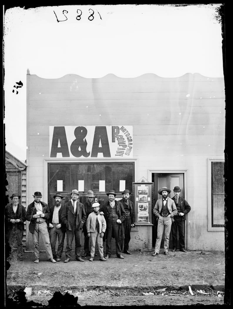 Group of smartly dressed men posing outside a shop with a sign above the window saying 'A&A Photographic Company'.