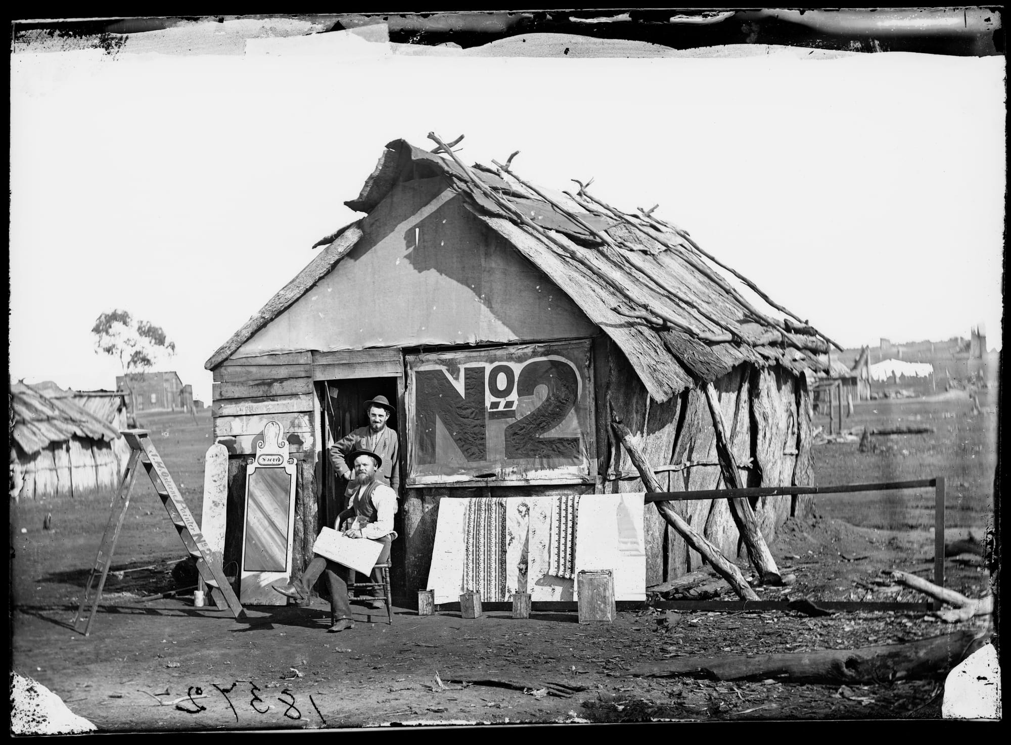 Two men posing outside a ramshakle timber shack with a large No.2 painted on the side, and bits of sign painting work around.