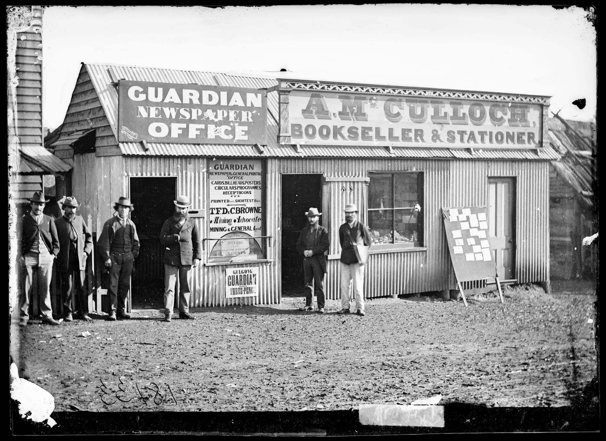 Smartly dressed me standing in front of a basic structure with signs advertising the Guardian Newspaper Office and A. McCulloch bookseller and stationer.