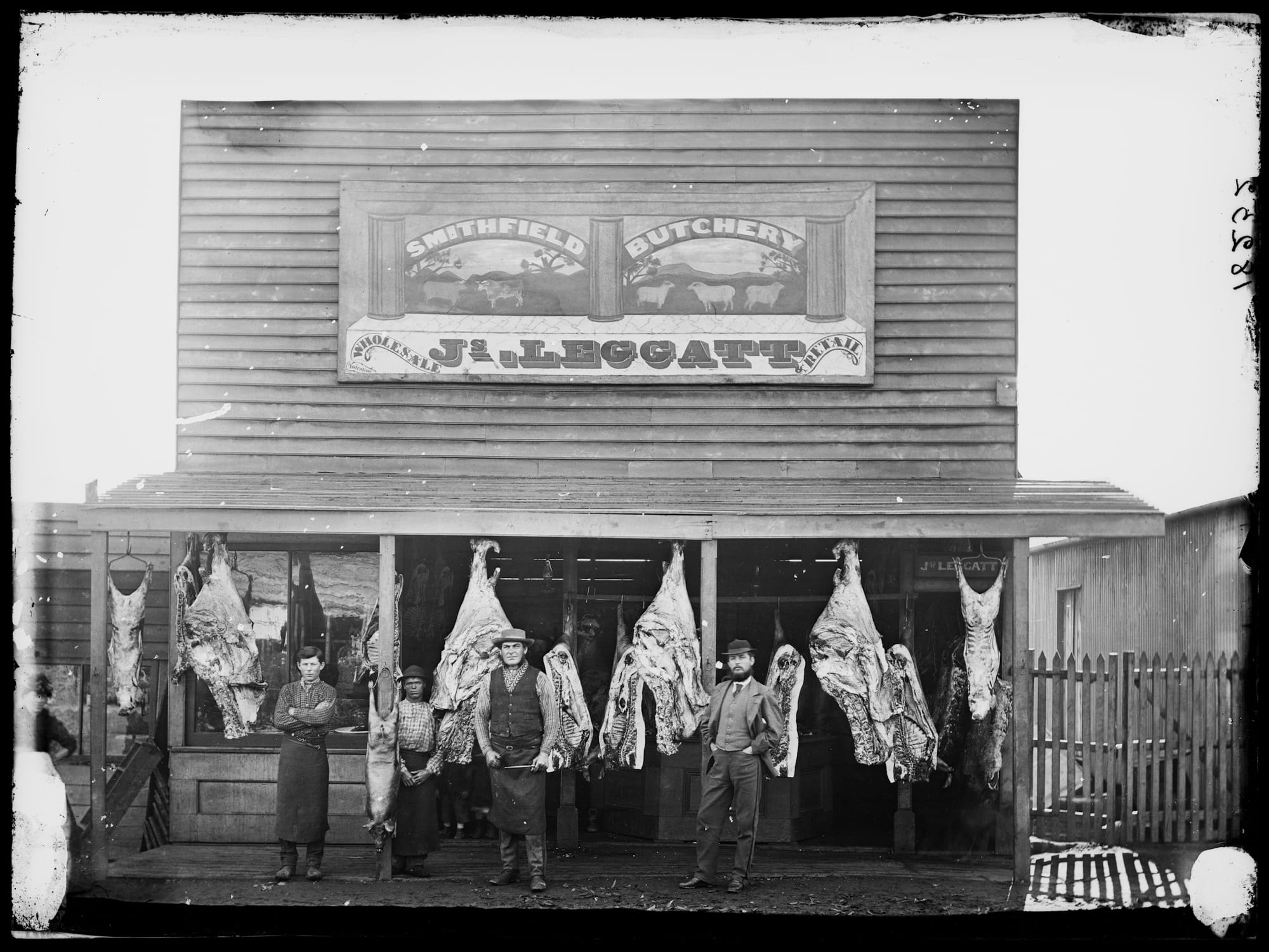 Three men standing in front of a butcher shop with carcasses hanging form the awning, and a sign mounted on the frontage above them.