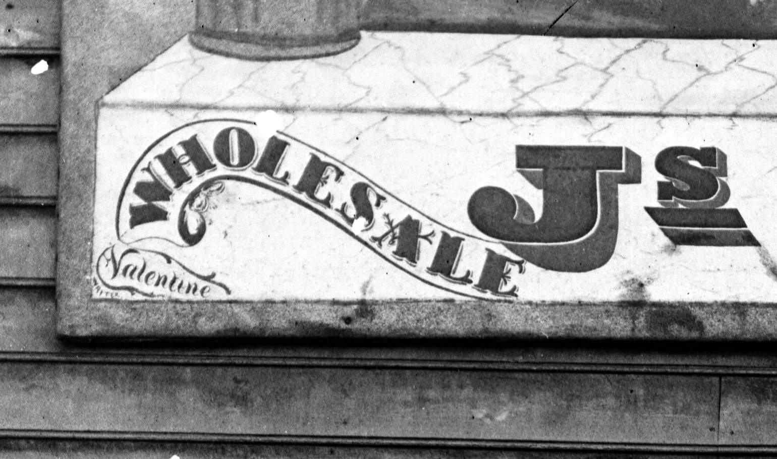 Corner of a mounted sign board showing a bit of the main letters ('Wholesale, Js') and the signature of the sign painter, Valentine bottom left.
