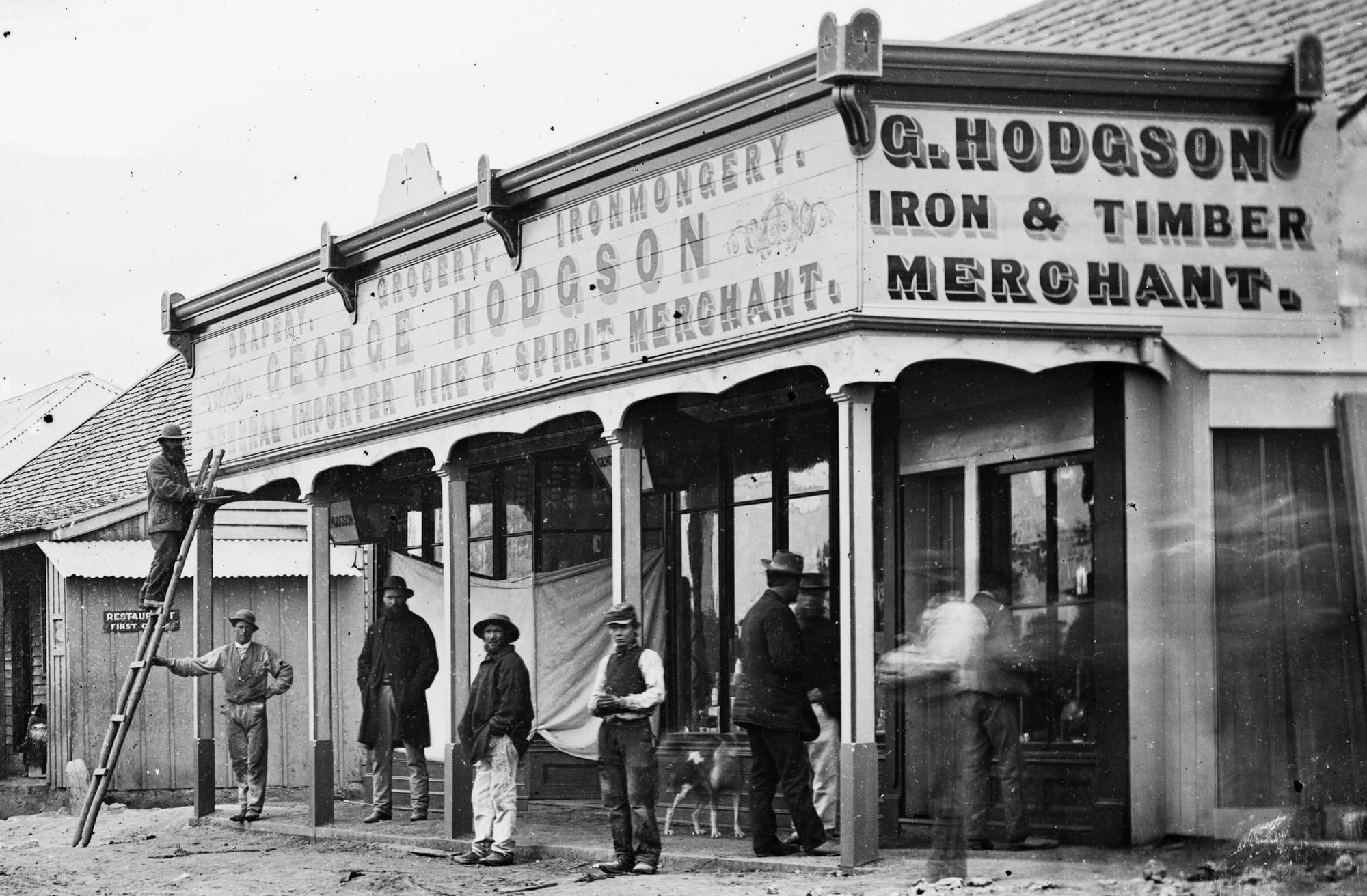 Large timber building housing George Hodgson's shop selling various goods. Men are posing in front, including one up a ladder painting a sign on the frontage.