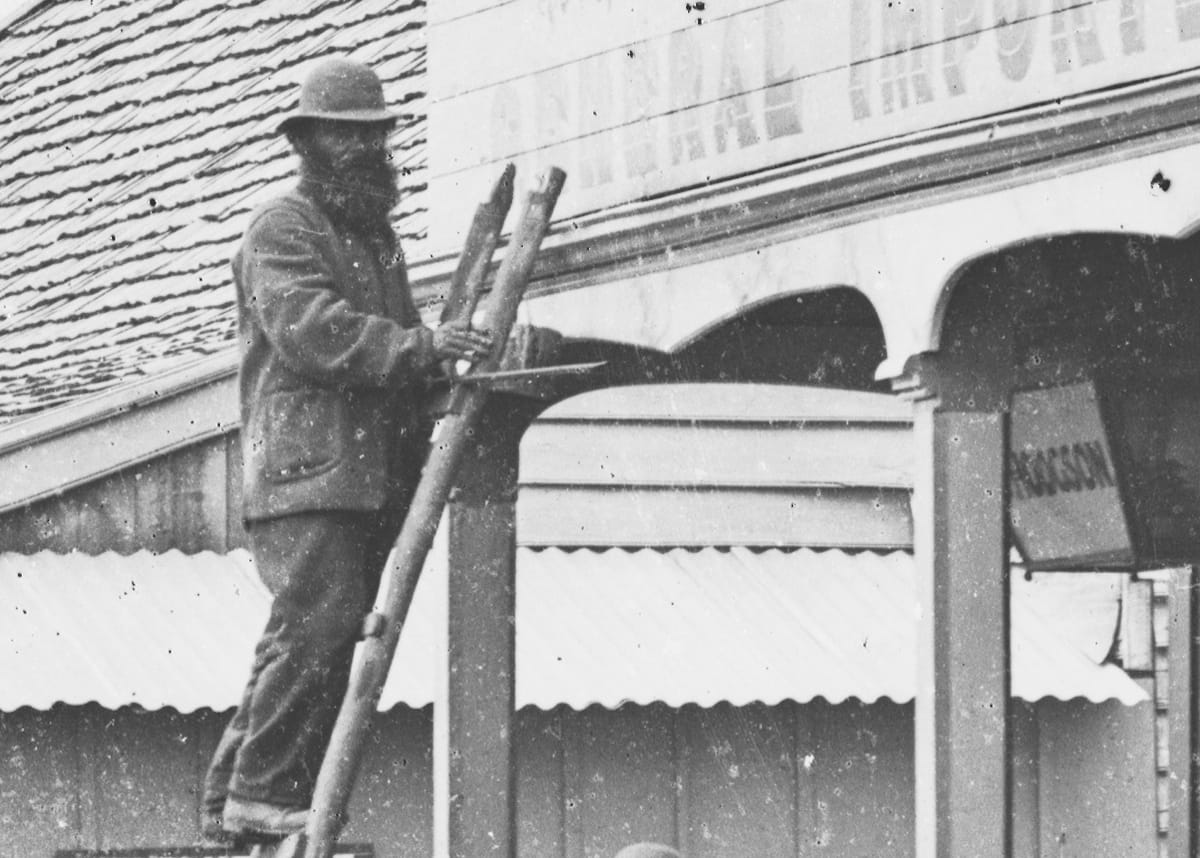 Man in a bowler hat on top of a ladder propped against a shopfront sign that he is working on.