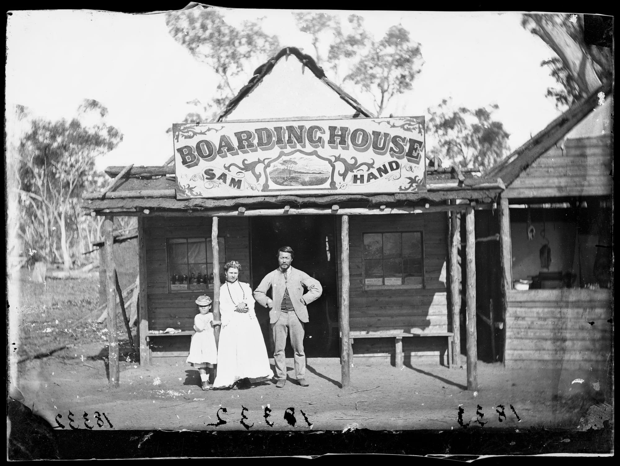 Family of three in smart clothes in front of a basic structure with a sign board for 'Boarding House, Sam Hand' mounted above the entranceway.