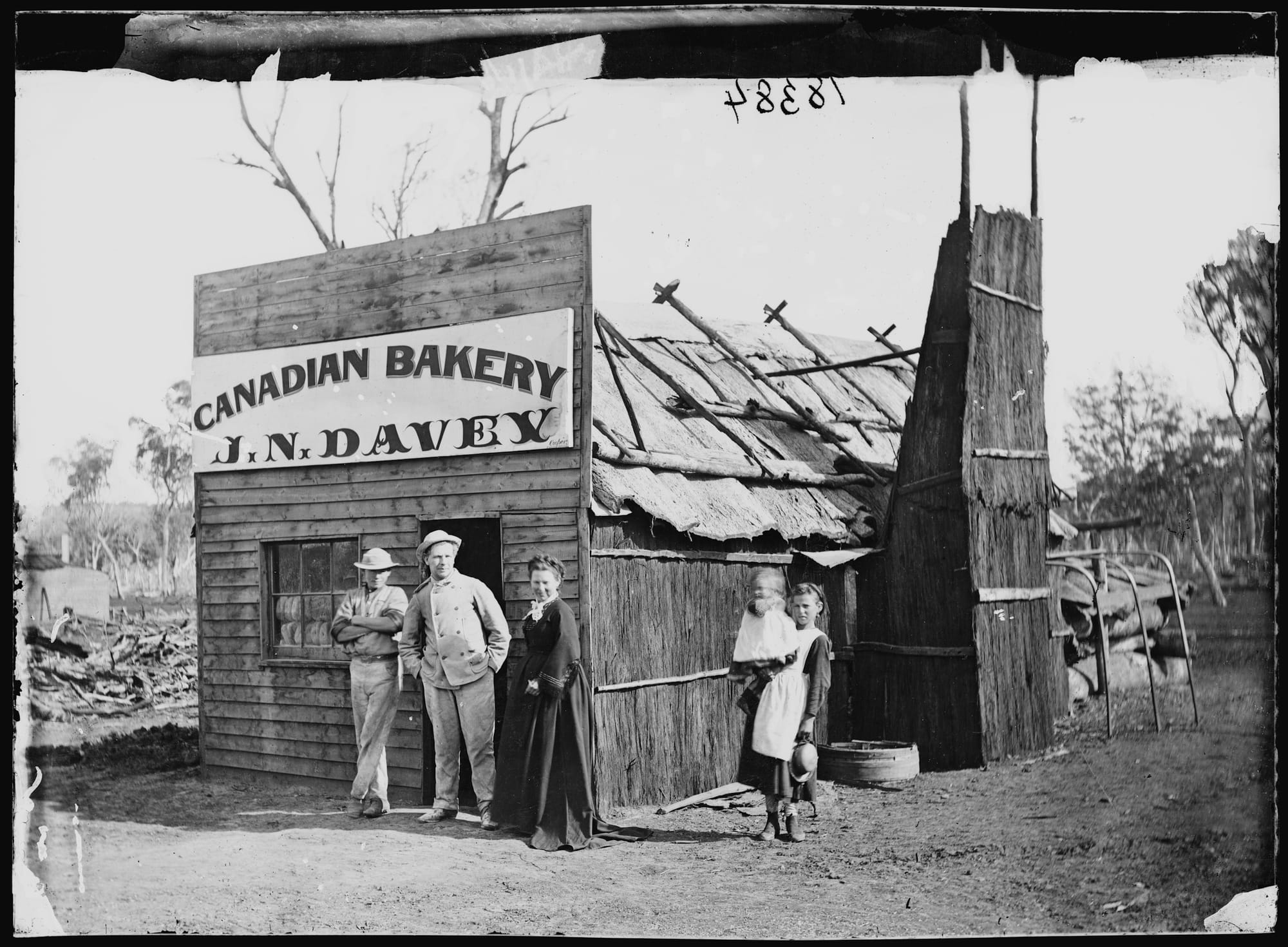 Four people and a baby posing in front of a basic structure that houses a bakery as advertised on the painted sign on the frontage.