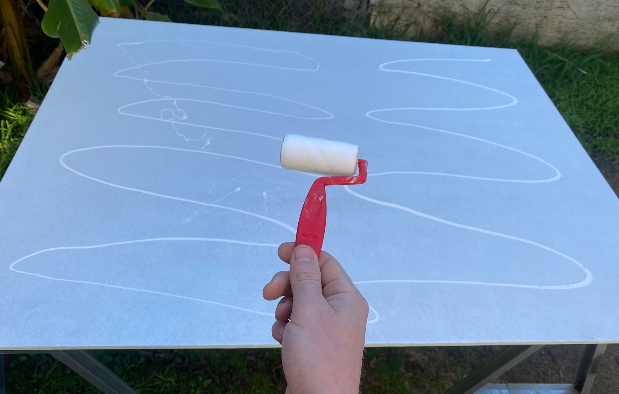 Basic paint roller being held in front of a flat board with zigzag lines of paint ready to be smoothed to an even finish.