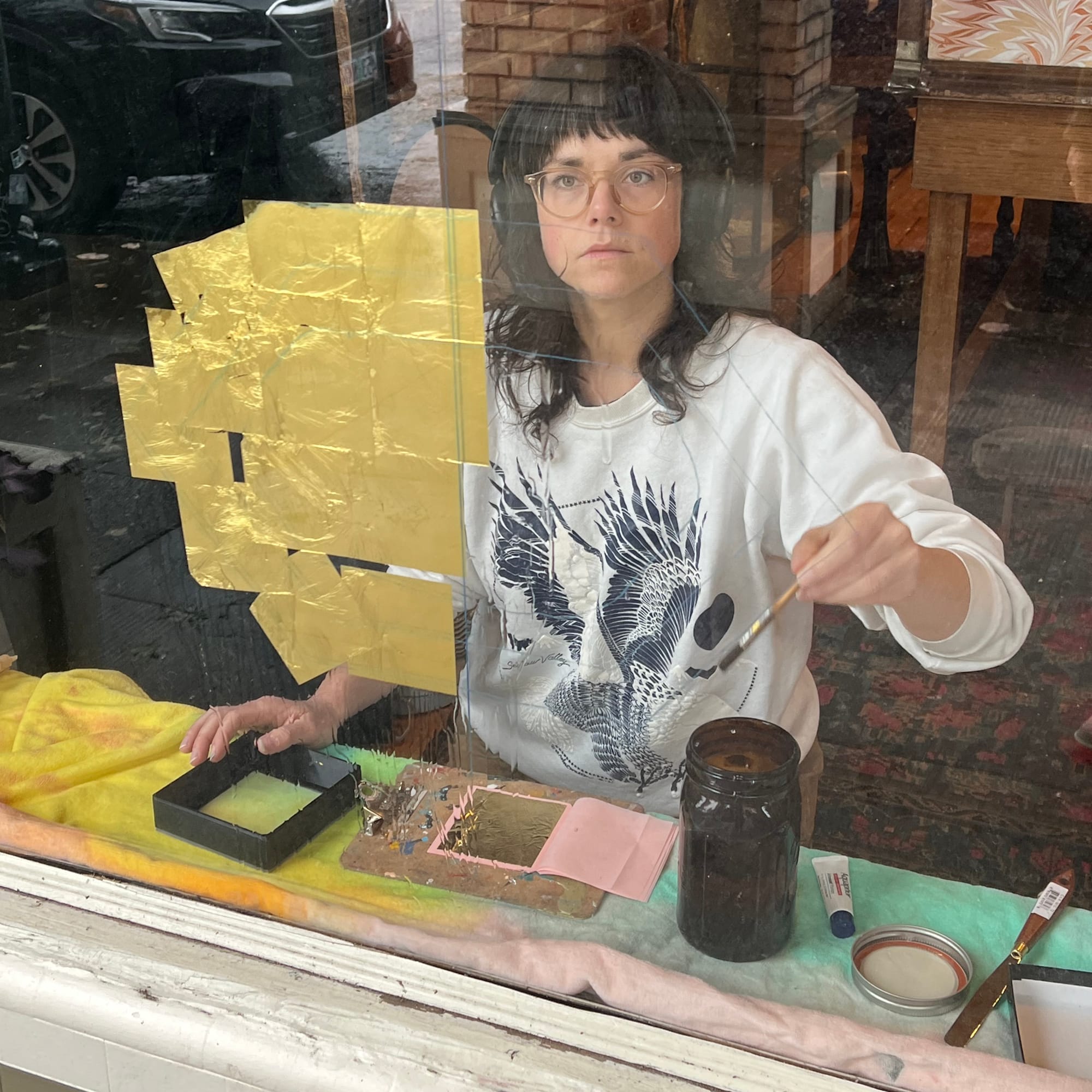 View through a window of a woman wearing headphones and in the process of places squares of gold on the glass.