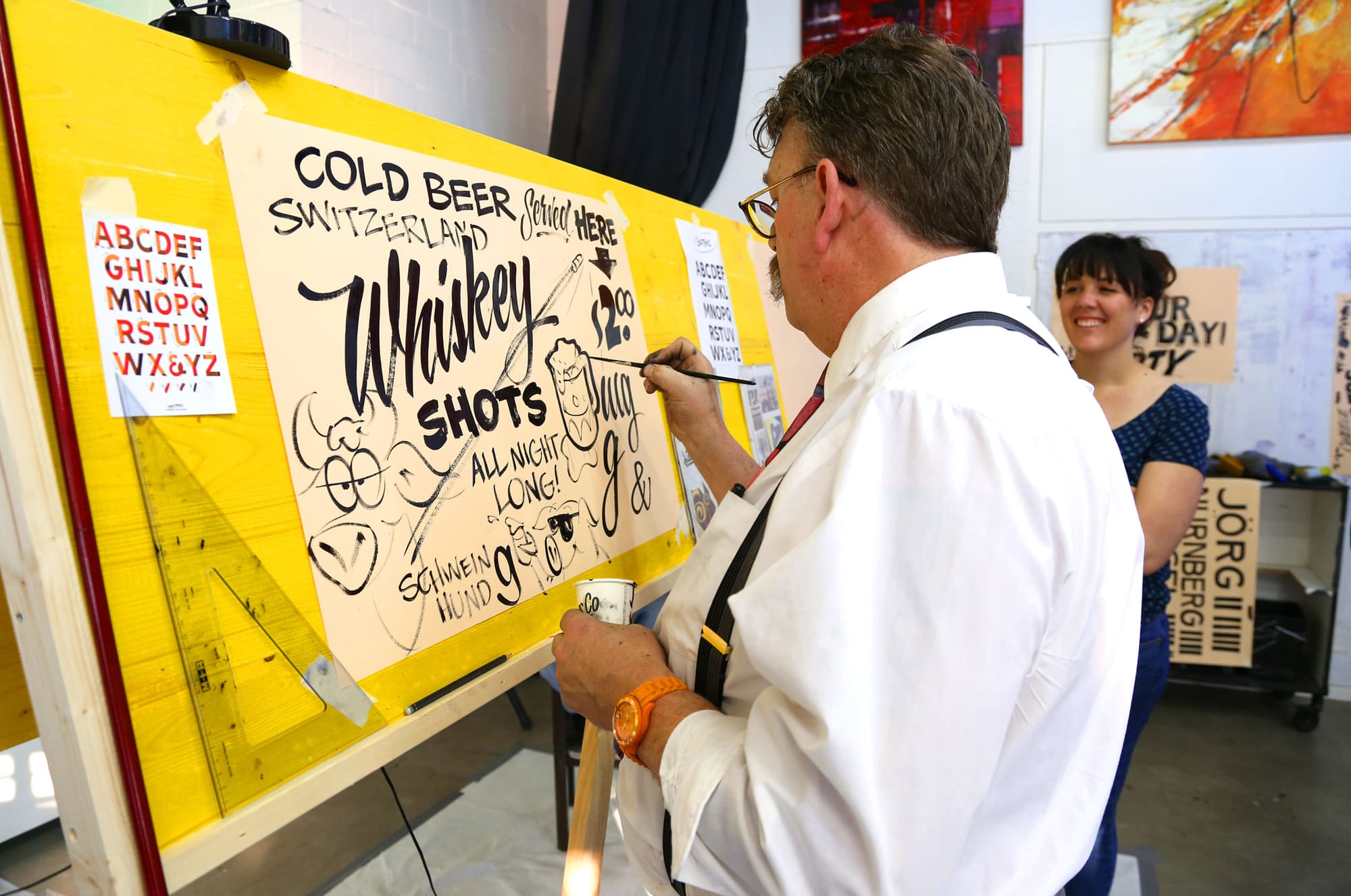 Man painting a sheet with different lettering styles and illustrations while a student looks on.