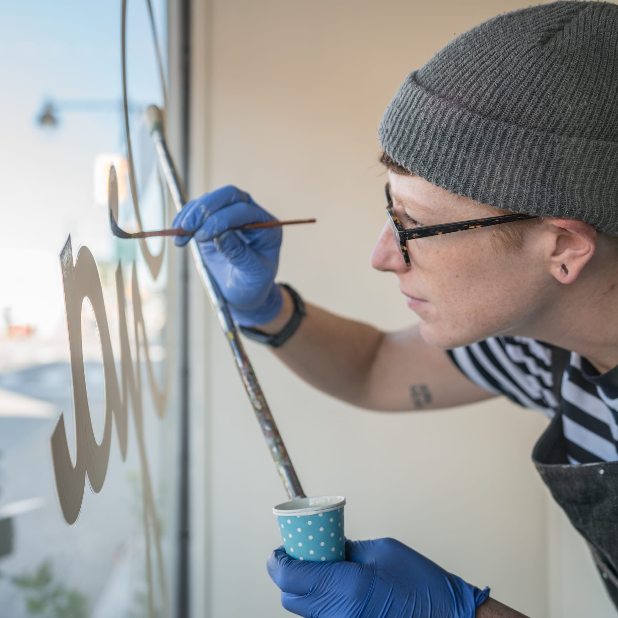 Woman in a hat and glasses concentrating as she paints white lettering onto a glass window.