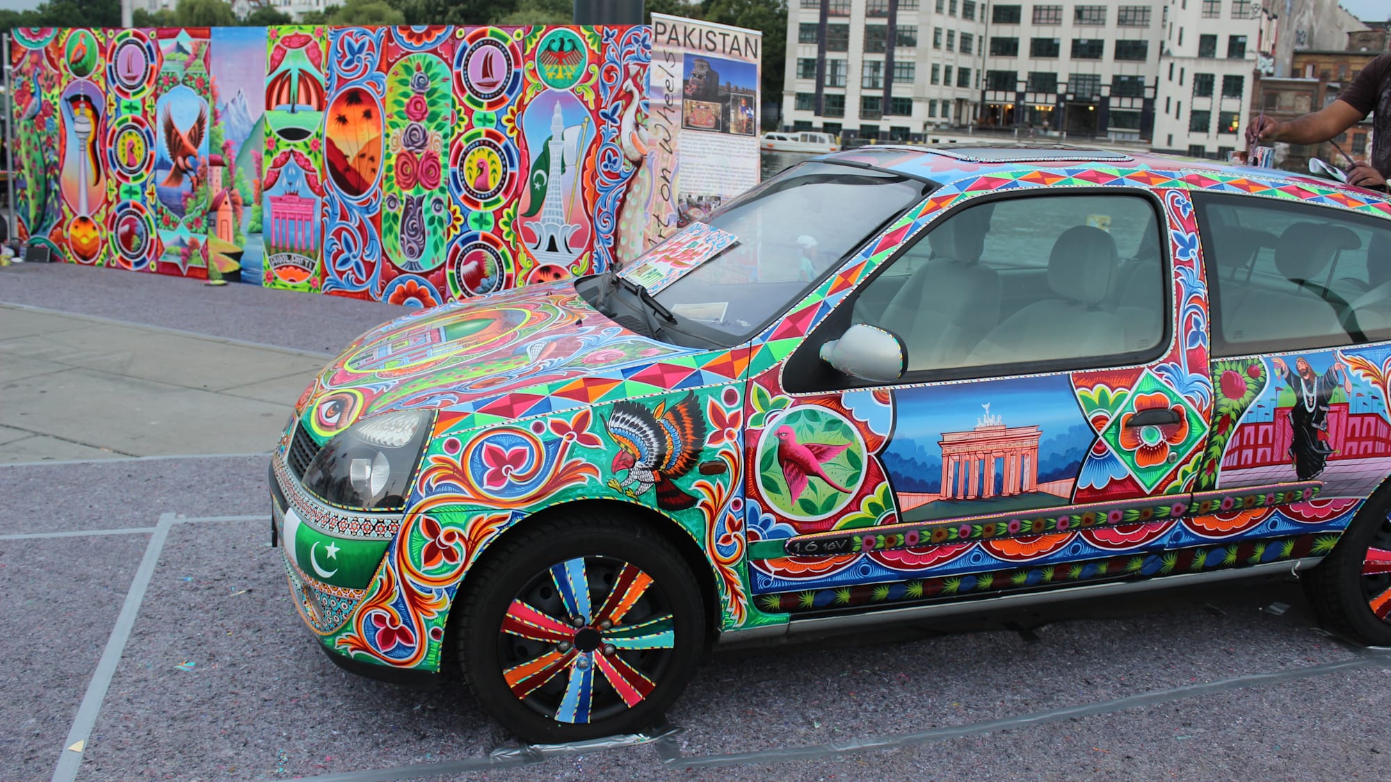 A car in front of a mural, with both decorated in brightly coloured pictorials, patterns and motifs.