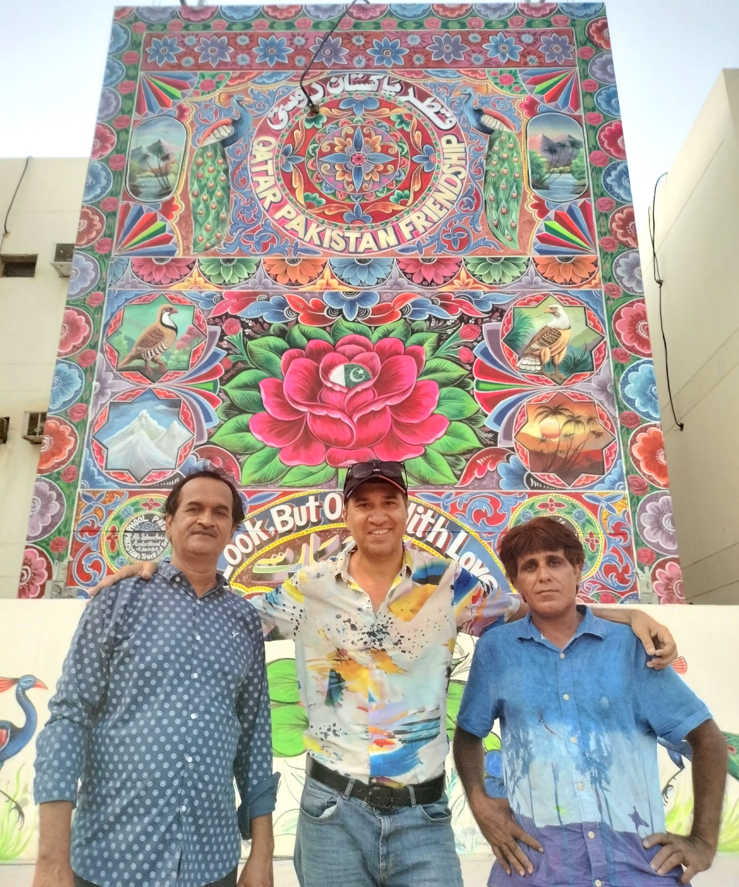 Three men standing in front of a very tall mural consisting of a brightly coloured design, and smaller lettering that reads "Qatar Pakistan Friendship".