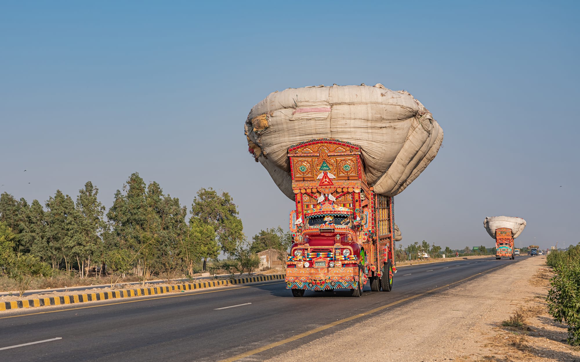Flamboyantly decorated truck carrying an enormous load as it makes its way along a road followed by a similarly decorated truck behind.