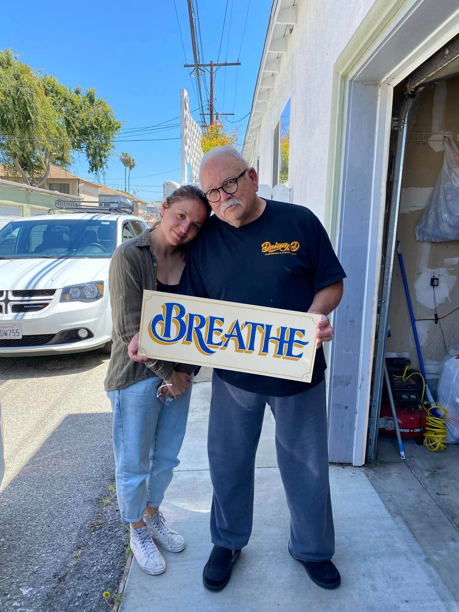 Man and woman posing in the shade on a sunny street holding a hand-painted sign that says 'Breathe'.