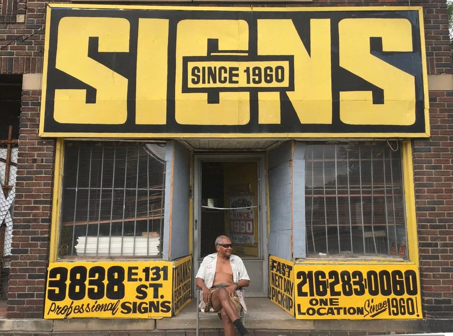 Man sitting in front of a shopfront which is emblazoned with a huge fascia sign that reads "SIGNS".