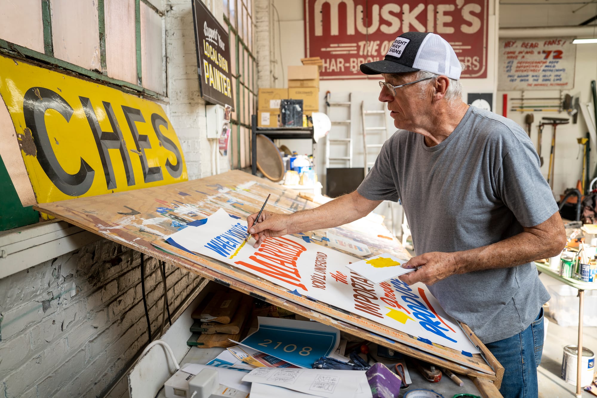 Man in a baseball cap working at an inclined easel painting a paper sign. The location is a sign shop with numerous pieces of signage around.