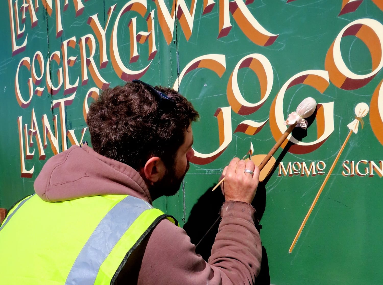 Sign painter painting with hand rested on a mahl stick. Behind is the nearly-finished sign in Roman lettering with a block shade.