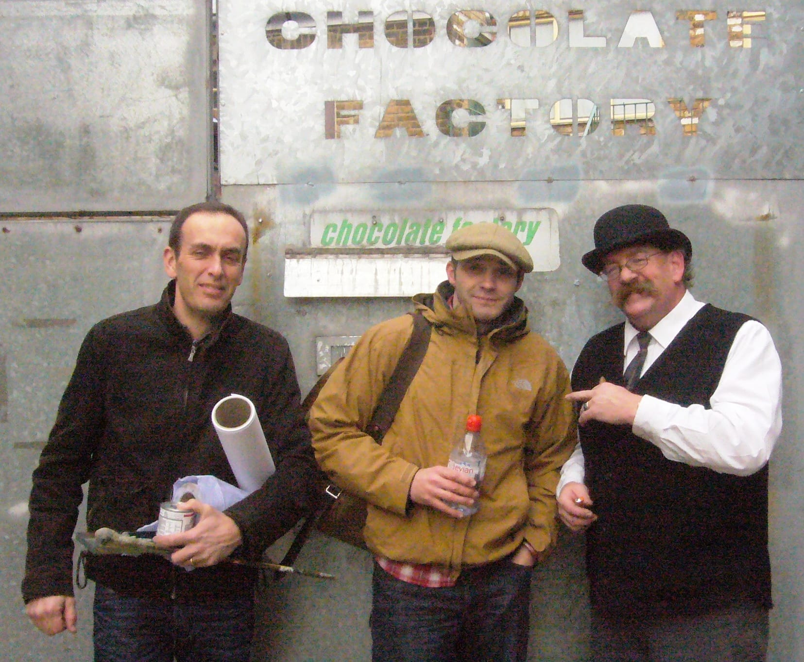 Three man standing in front of a metal gate with the words "Chocolate Factory" cut out above their heads.