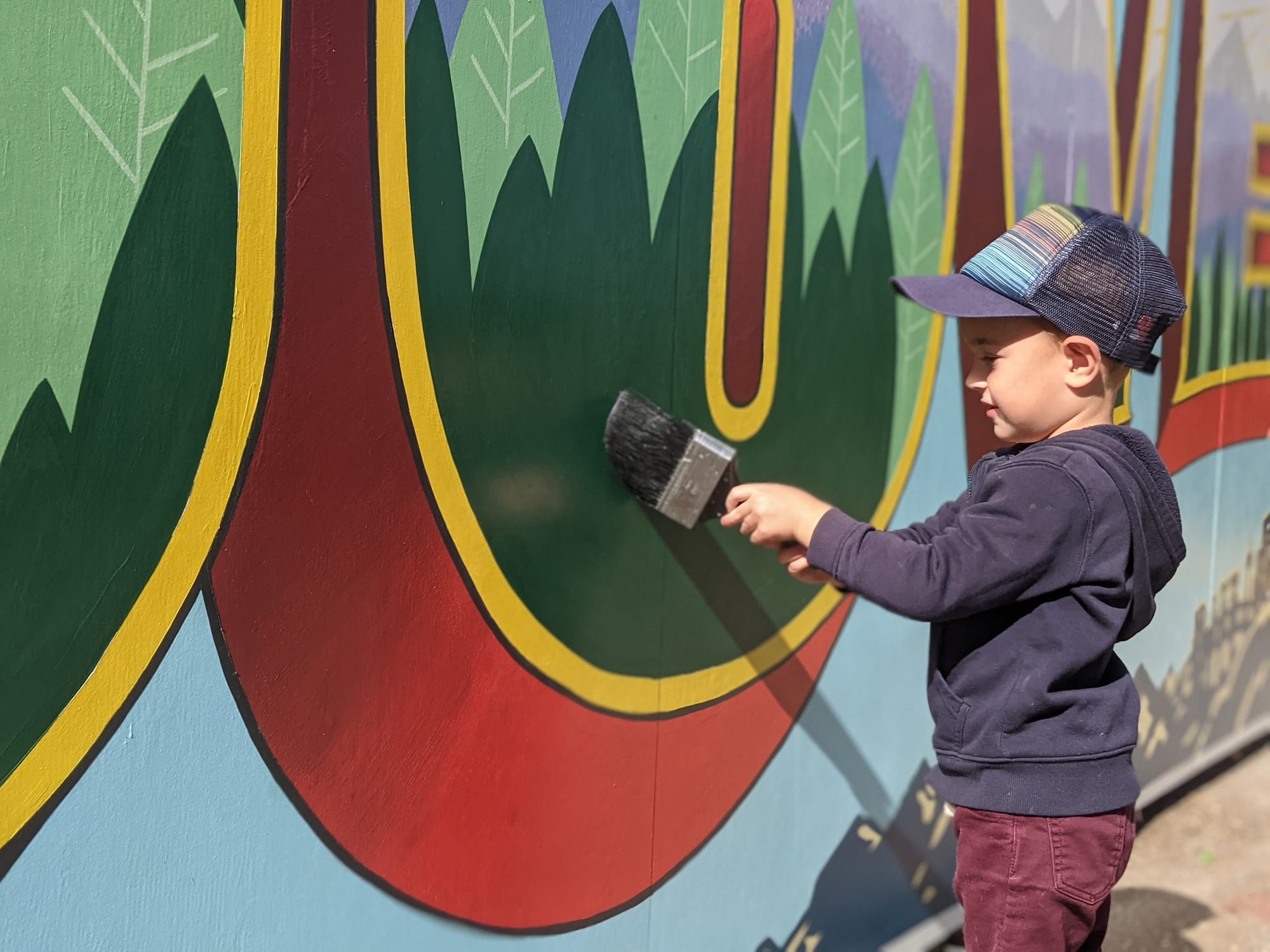 Young boy in a baseball cap painting a giant letter on a wall with a huge brush.